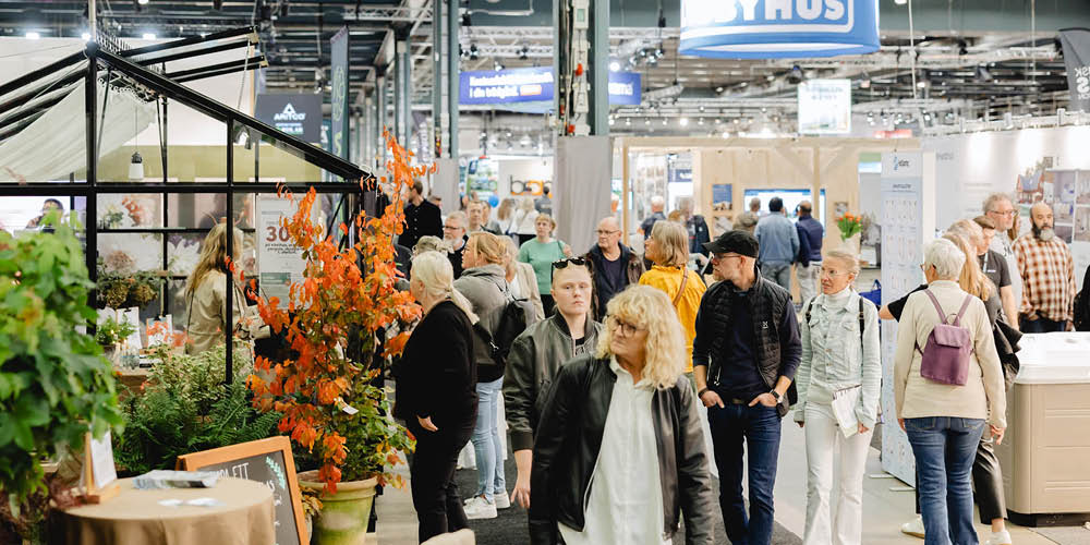 HVM_upptack_1000x500px_NY Vy över en mässgång på Hem & Villamässan. Människor strosar runt och tittar. I bilden syns ett svart växthus och en stor växt med orange blad.